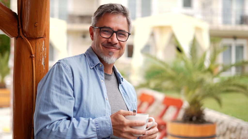 Portrait of happy middle aged man at home outdoor on terrace in garden, relaxing, drinking morning coffee, smiling.